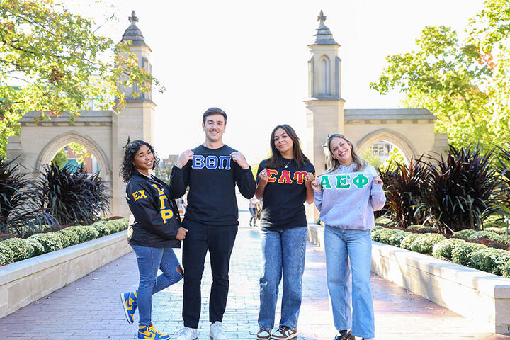 Four students wearing Greek letter sweatshirts stand in front of the Sample Gates.